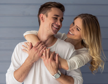 Portrait of beautiful young couple in casual clothes hugging, looking at each other and smiling, standing against gray wallの写真素材