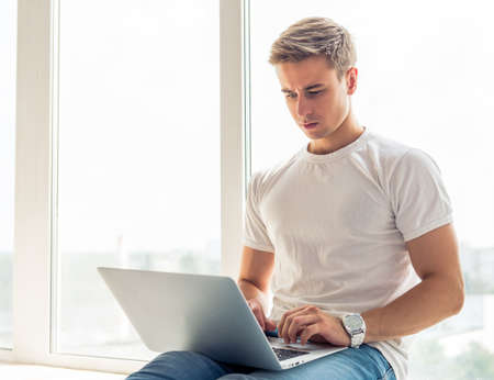 Handsome young businessman in casual clothes is using a laptop and working while sitting at home before the windowsの写真素材