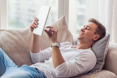 Handsome young businessman in casual clothes is using a digital tablet and smiling while lying on...