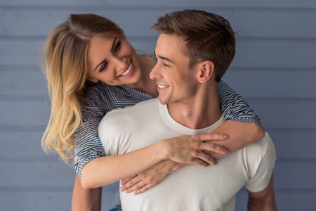 Portrait of beautiful young couple in casual clothes hugging, looking at camera and smiling, standing against gray wallの写真素材