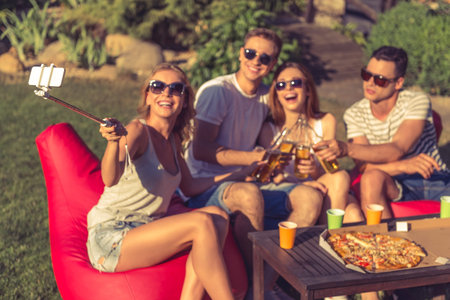 Young girl is making selfie with her friends who are clinking bottles of beverage and smiling, sitting on bean bag chairs outdoors. Phone and monopod in focusの写真素材