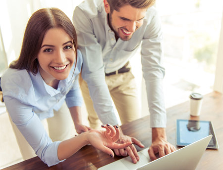 Beautiful young business woman and handsome businessman are using a laptop, talking and smiling...