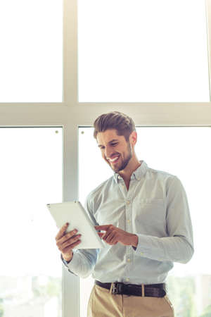 Handsome young businessman in classic shirt is using a digital tablet and smiling, standing in office before the windowの写真素材