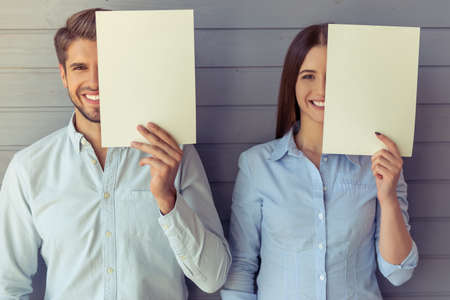 Beautiful young business woman and businessman are holding white sheets of paper and covering half of their faces, looking at camera and smiling, standing before gray wallの写真素材