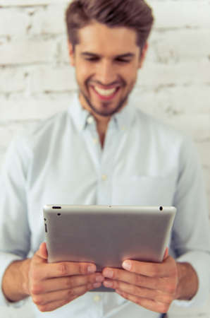 Handsome young businessman in classic shirt is using a digital tablet and smiling, standing in front of white brick wallの写真素材