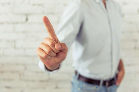 Cropped image of handsome young businessman in classic shirt keeping his finger up, standing in front of white brick wall. Hand in focusの写真素材