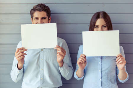 Beautiful young business woman and businessman are holding white sheets of paper and covering half of their faces, looking at camera and smiling, standing before gray wallの写真素材