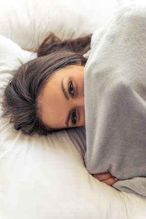 Portrait of beautiful young woman looking at camera and hiding behind blanket while lying in her bed at homeの写真素材