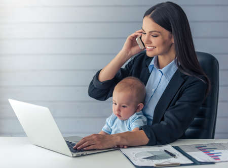 Beautiful business lady in classic suit is talking on the mobile phone and smiling, working in office while her cute little baby is sitting on her kneesの写真素材