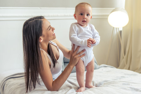Beautiful young mom and her sweet little baby are smiling while spending time together on bed at...