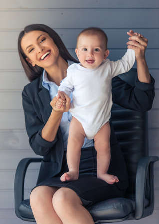 Portrait of beautiful business lady in classic suit and her sweet little baby looking at camera and smiling, sitting on office chair on gray backgroundの写真素材