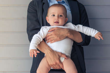 Portrait of sweet little baby looking at camera and smiling while mom is holding him, standing before gray wall, croppedの写真素材