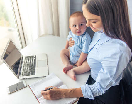 High angle view of beautiful business lady holding her cute little baby, making notes and smiling while working in office. Baby is looking on his momの写真素材