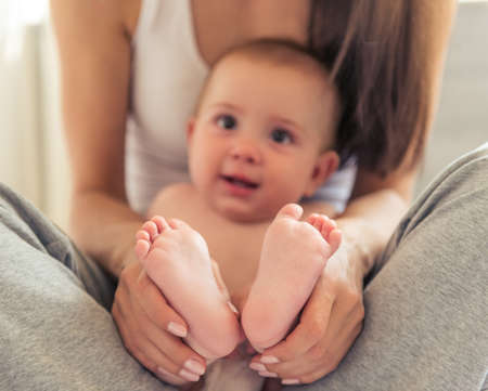 Portrait of charming naked little baby looking away and smiling while sitting on his mother's knees. Baby's bare feet in focusの写真素材