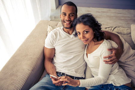Beautiful young Afro American couple is using a smartphone, looking at camera and smiling while...