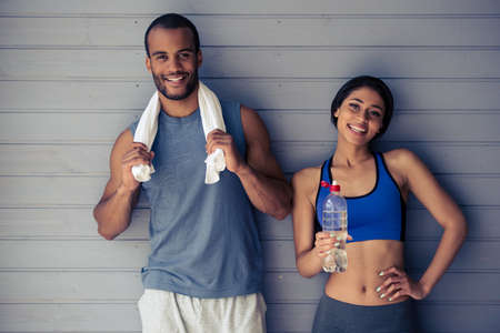 Beautiful Afro American couple in sports clothes is holding a bottle of water and a towel, looking at camera and smiling, standing in front of gray wallの写真素材