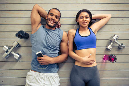 Top view of beautiful Afro American couple in sports clothes and earphones listening to music, looking at camera and smiling while resting on the floor after workoutの写真素材