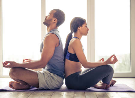 Beautiful Afro American couple in sports clothes is meditating sitting back to back in lotus position on mat while doing yoga at homeの写真素材