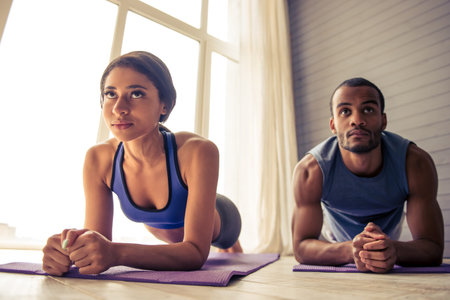Beautiful Afro American couple in sports clothes is doing plank on yoga mat while working out at...