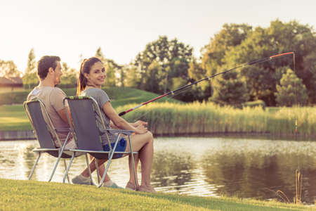 Back view of beautiful couple catching fish in the pond using a fishing rod. Girl is looking at camera and smilingの写真素材