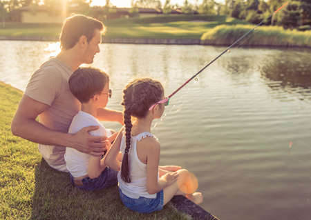 Back view of handsome father and his beautiful little kids catching fish in the pond using a fishing rod, sitting on the groundの写真素材