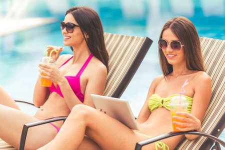 Beautiful girls in swimwear and sun glasses are drinking juice, using a digital tablet and smiling while sunbathing on the chaise longue near the poolの写真素材