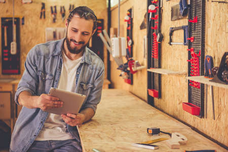 Handsome carpenter is using a digital tablet and smiling while working in the workshopの写真素材