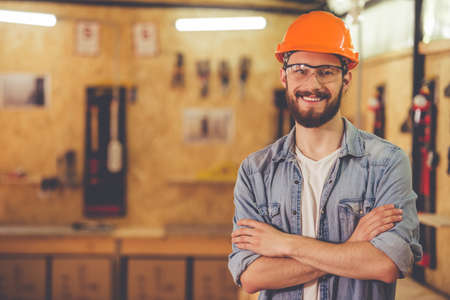 Handsome carpenter in hard hat and protective glasses is looking at camera and smiling while standing with crossed arms in the workshopの写真素材