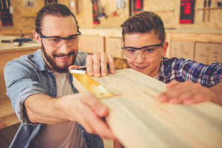 Handsome teenage carpenter and his father in protective glasses are measuring wood and smiling while working in the workshopの写真素材