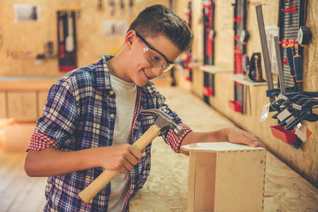 Handsome teenage carpenter in protective glasses is smiling while working with wood and a hammer in the workshopの写真素材