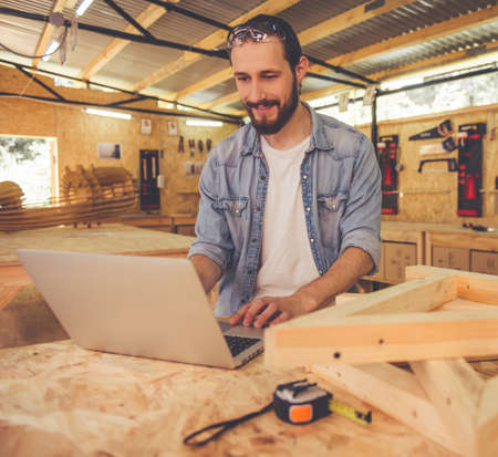 Handsome carpenter is using a laptop and smiling while working in the workshopの写真素材