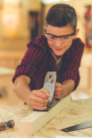 Handsome teenage carpenter in protective glasses is smiling while working with wood and carpenter's plane in the studioの写真素材