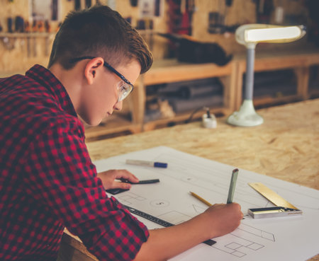 Handsome teenage carpenter in protective glasses is drawing his new project in the workshop