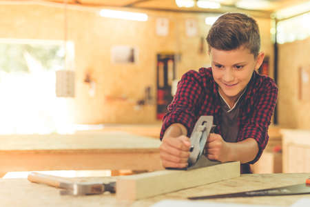 Handsome teenage carpenter is smiling while working with wood and carpenter's plane in the studioの写真素材