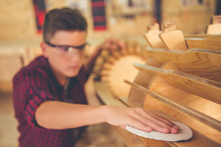 Handsome teenage carpenter in protective glasses is grinding wood using sandpaper in the workshop. Hand in focusの写真素材