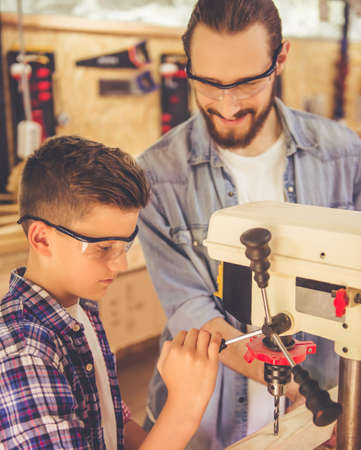 Handsome carpenter and his son in protective glasses are smiling while working with wood and drilling machine in the workshopの写真素材