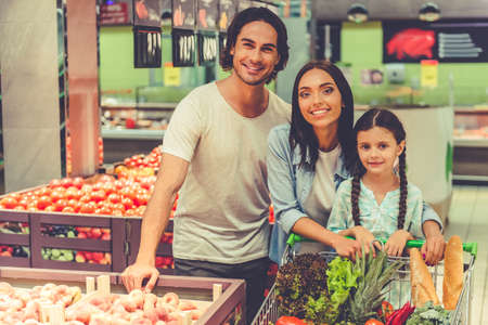 Beautiful young parents and their cute little daughter are looking at camera and smiling while choosing food in the supermarketの写真素材