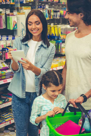 Beautiful young parents and their cute little daughter are smiling while choosing school stationery in the supermarket. Mom is making notes in the listの写真素材