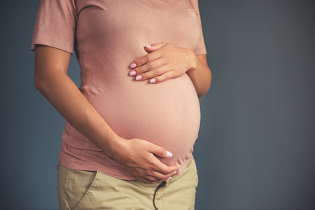 Cropped image of beautiful pregnant young woman in casual clothes touching her tummy, on a gray...