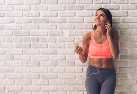 Beautiful young sportswoman in sportswear and earphones is listening to music using a smartphone, looking upward and smiling, on white brick wall backgroundの写真素材