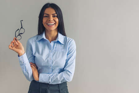 Beautiful young girl in smart casual wear is holding eyeglasses, looking at camera and smiling, on gray backgroundの写真素材