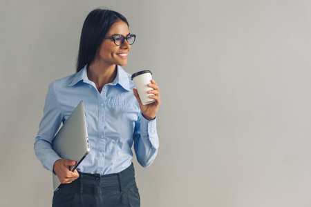 Beautiful young girl in smart casual wear and eyeglasses is holding a laptop, drinking coffee, looking away and smiling, on gray backgroundの写真素材