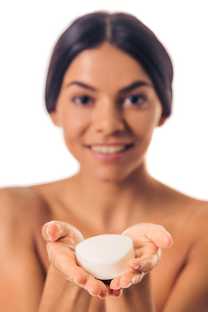 Portrait of beautiful girl with bare shoulders holding a soap, looking at camera and smiling, isolated on white. Hands with soap in focusの写真素材