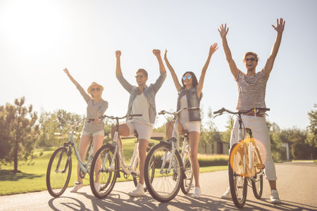 Beautiful stylish friends are holding hands up and smiling while cycling in the park