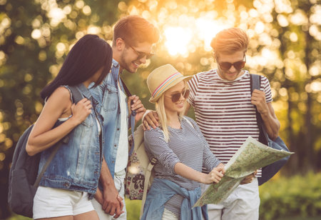 Beautiful stylish friends with backpacks are examining map and smiling while travelingの写真素材