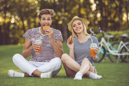 Beautiful stylish couple is drinking cocktails, eating sandwiches, talking and smiling while resting in the parkの写真素材