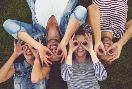Top view of beautiful stylish friends making glasses of fingers, looking at camera and smiling while lying on grassの写真素材