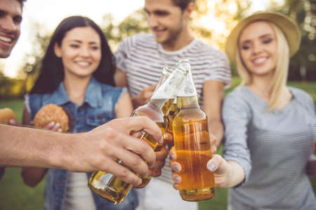 Cropped image of stylish friends clinking bottles of beverage, eating sandwiches and smiling while resting in the parkの写真素材