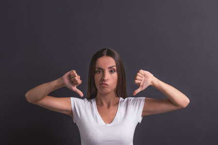 Beautiful young girl in white t-shirt is showing thumbs down and looking at camera, standing against blackboardの写真素材