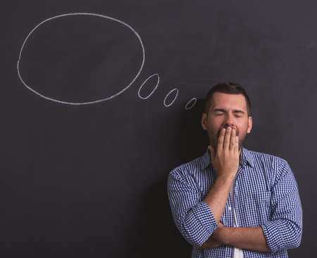 Handsome young tired man is covering his mouth while yawning, standing against blackboard with drawn speech bubbleの写真素材